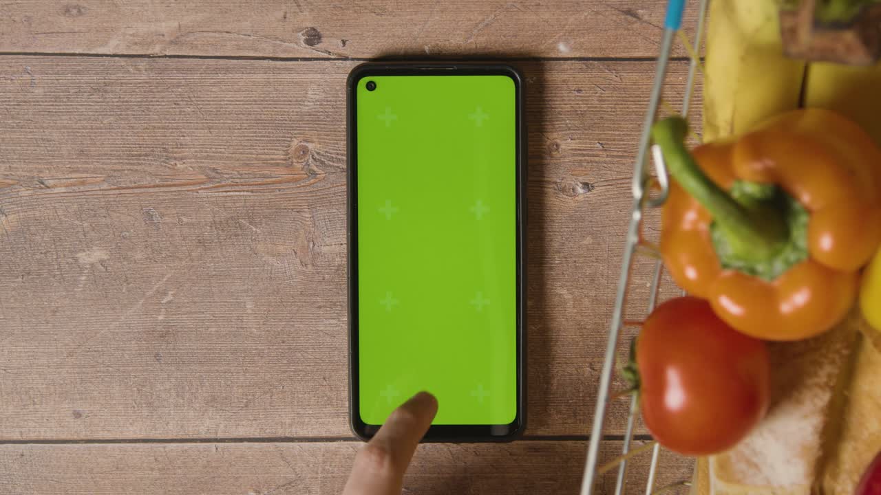 foto de estudio de una persona que usa un teléfono móvil de pantalla verde junto a alimentos básicos en la cesta de la compra de alambre del supermercado 1