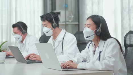 three asian doctors with stethoscopes in headsets and masks working as call center agents sitting on the chairs and typing while speaking to customers on the call at the office
