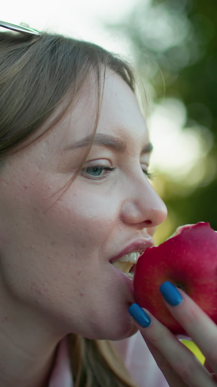 Close up of lady smiling softly while taking a gentle bite from a red apple, with a beautifully blurred green background, enhancing a moment of relaxation and enjoyment in nature