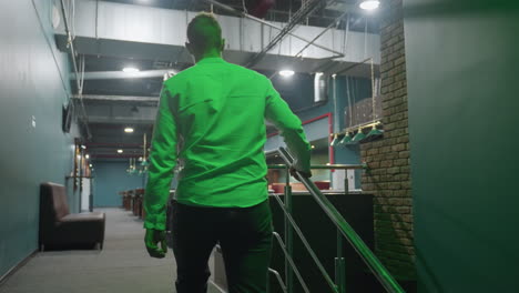 back shot of young man in shirt and trousers walking on stairs, holding rail while heading towards billiard room. green lighting adds atmospheric effect with stylish industrial design