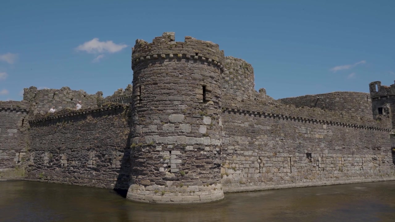 Vista del Castello di Beaumaris con il suo ampio fossato chiaramente visibile in primo piano, che mostra acqua calma, mura e torri in pietra consumate dalle intemperie circondate da erba verde lussureggiante e campagna naturale