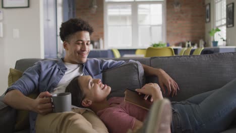 happy biracial couple sitting on sofa in living room, drinking coffee