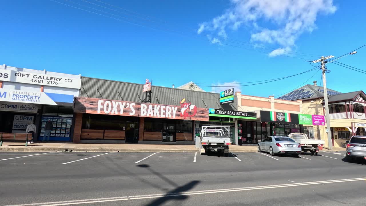 A vehicle travels along a quiet urban street lined with local shops, parked cars, and clear blue skies, captured in smooth daylight motion