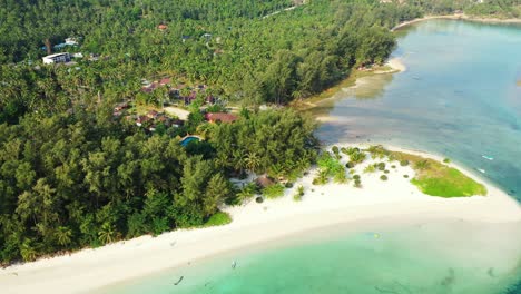 Holiday-resort-with-bungalows-inside-tropical-trees-forest,-white-sandy-beach-washed-by-calm-clear-water-of-lagoon-in-Thailand