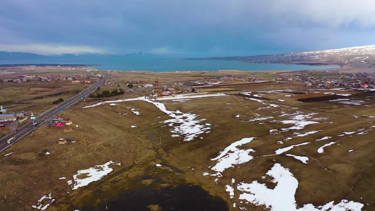 Ländliches Dorf mit Autobahn und Häusern, Drohnenansicht von türkisfarbenem Wasser, fleckigem Schnee und dramatischen Wolken.