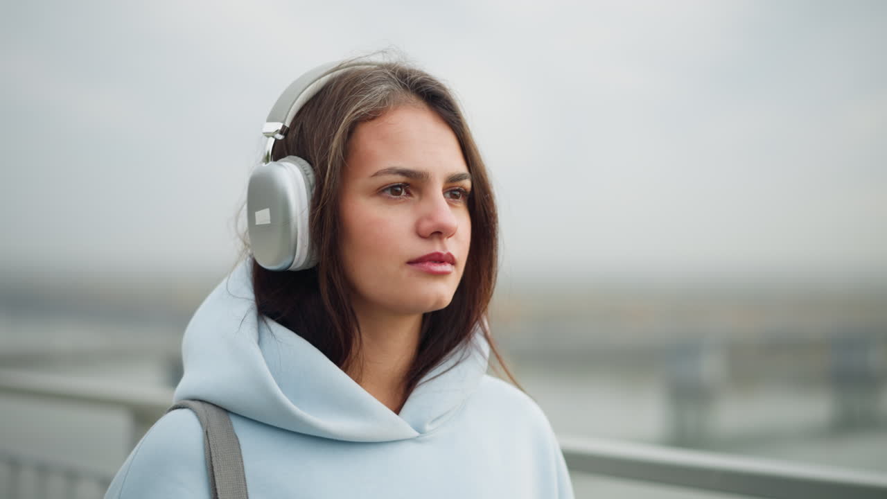 Close-up view of lady in casual wear listening to music with headphones, focused expression, blurred background featuring bridge and flowing water, tranquil setting outdoors