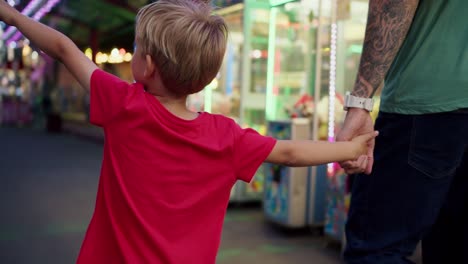 un niño pequeño feliz rubio en una camiseta roja con su padre ir y mirar los paseos en el parque de atracciones. un niño pequeño en una camisa roja y su padre están activamente mirando los paseos en el parque de diversiones mientras camina a lo largo de ellos