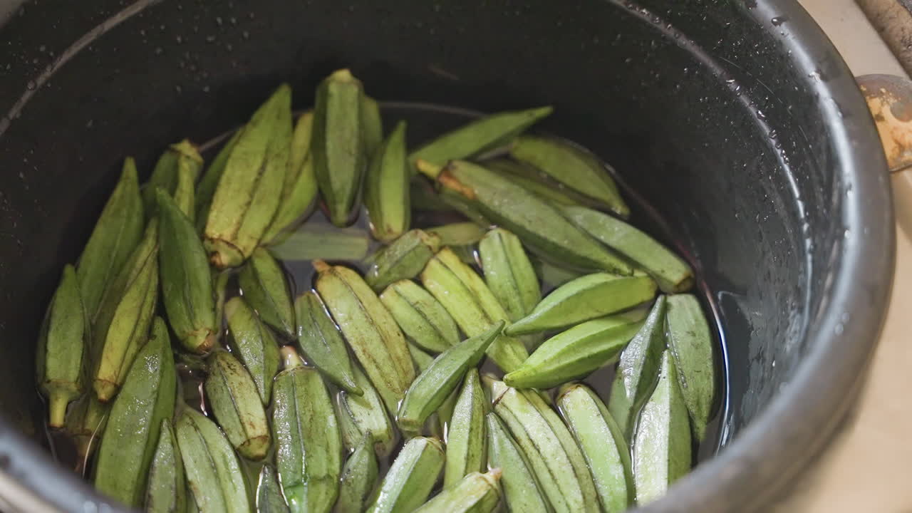 Okra segar direndam dalam panci, air berkilauan, tampilan close-up dari atas, adegan persiapan dapur menunjukkan polong hijau bertumpuk dalam panci berbintik, pantulan cahaya, tekstur basah, persiapan untuk memasak, rumah alami
