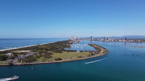 high fast moving drone view travelling over a man-made ocean inlet towards an urban boat harbor with a metropolitan skyline and mountain range in the distance