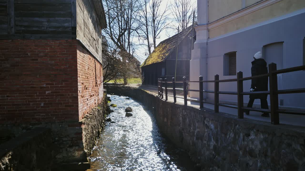 Bright stream flows between vintage houses as woman enjoys peaceful spring walk