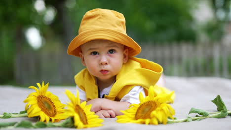lovely serious kid in yellow panama and vest lies on his belly outdoors. sunflowers lie in front of a kid. blurred nature backdrop.