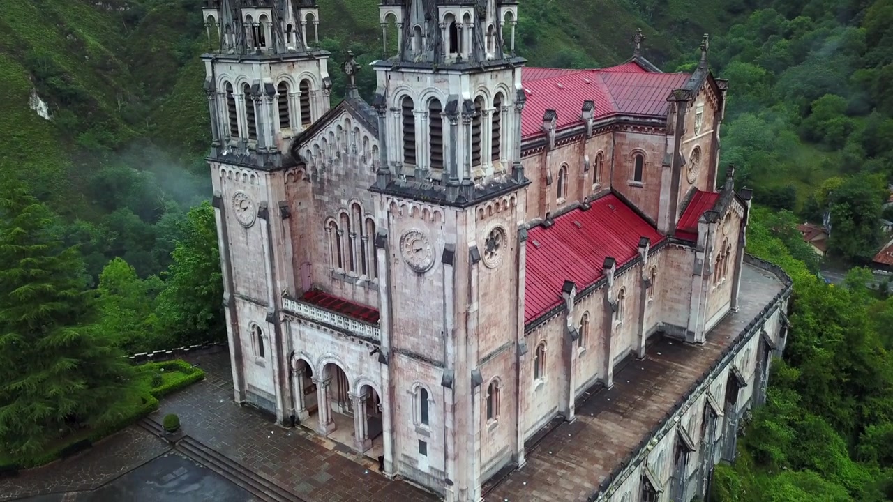 Aerial drone footage of the Covadonga basilica and Santa Cueva sacred cave shrine in Asturias Spain. Gothic towers and green mountain landscape surround this Marian pilgrimage site with Don Pelayo