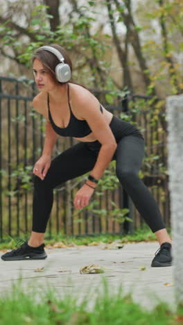 young girl in black activewear stretching her legs outdoors, tapping her toe with hand while listening to music, blurred background showing iron fence and trees, enjoying fitness routine