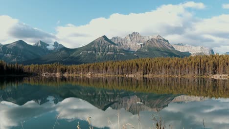 Un-Hermoso-Lapso-De-Tiempo-De-Una-Montaña-En-Algún-Lugar-De-Banff,-Con-Un-Lago-Y-El-Reflejo-De-Las-Montañas-Que-Se-Muestran-En-Las-Tranquilas-Aguas-Del-Lago,-Con-Nubes-Que-Pasan-Y-El-Sol-Asomándose-A-Través-De-Ellas