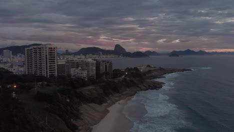Luftannäherung-An-Einen-Kleinen-Strand-Mit-Der-Copacabana-Festung-Im-Vordergrund-Und-Dem-Zuckerhut-Im-Hintergrund-Vor-Einem-Bewölkten-Sonnenaufgang