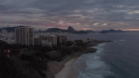 Enfoque-Aéreo-De-Una-Pequeña-Playa-Con-El-Fuerte-De-Copacabana-En-Primer-Plano-Y-La-Montaña-Sugarloaf-En-El-Fondo-Contra-Un-Amanecer-De-Color-Nublado