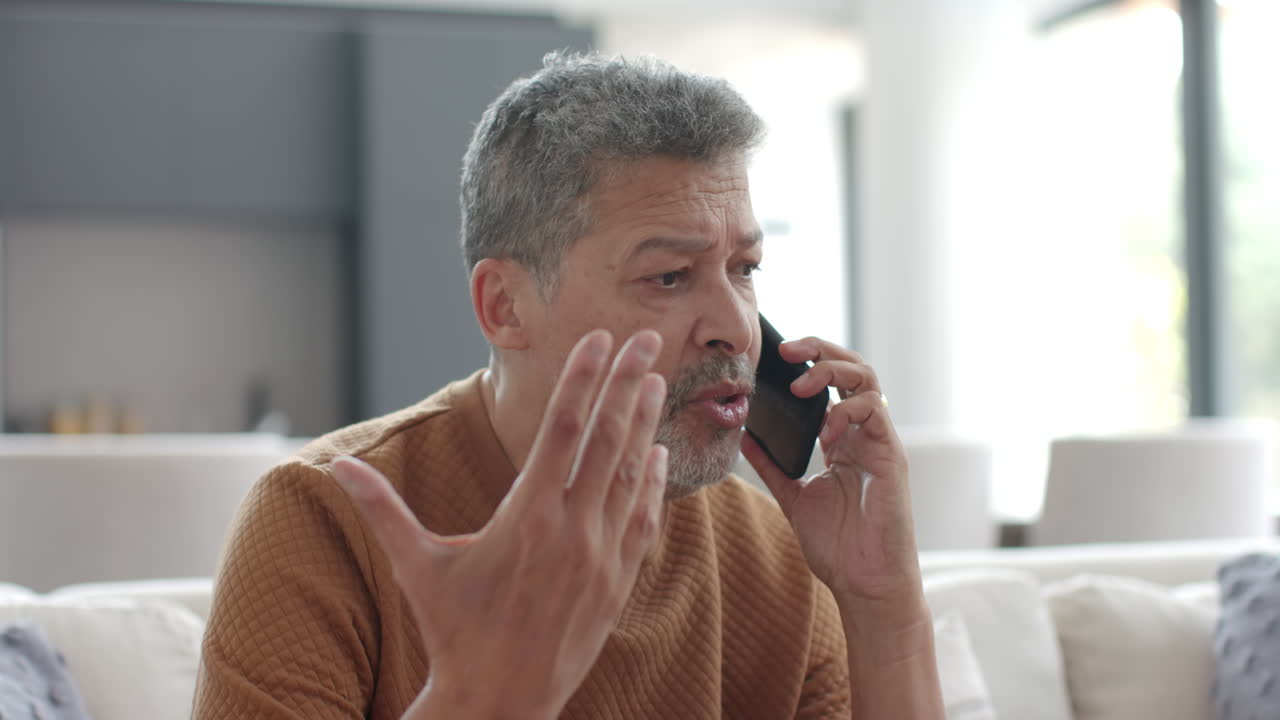 Senior biracial man talking on smartphone sitting on couch in living room, slow motion