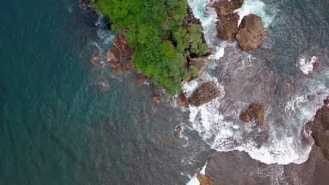 Toma-Aérea-De-Arriba-Hacia-Abajo-Del-Agua-Que-Choca-Contra-Rocas-Y-Cantos-Rodados-Cerca-De-La-Playa-De-Jungwok,-Indonesia