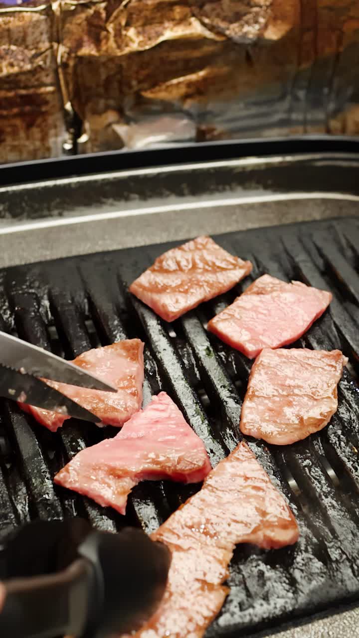 Bidikan jarak dekat tangan yang menggunakan gunting dan penjepit untuk memotong daging sapi di atas panggangan arang