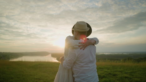 two lovers embrace warmly in a grassy field at sunset, both wearing white outfits and hats. the golden light of the setting sun enhances the romantic atmosphere as they hold each other close