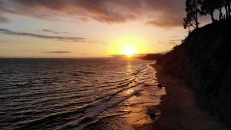 toma aérea de drones de un cielo de puesta de sol naranja sobre las olas del océano frente a los acantilados de la playa de arena de santa bárbara, california