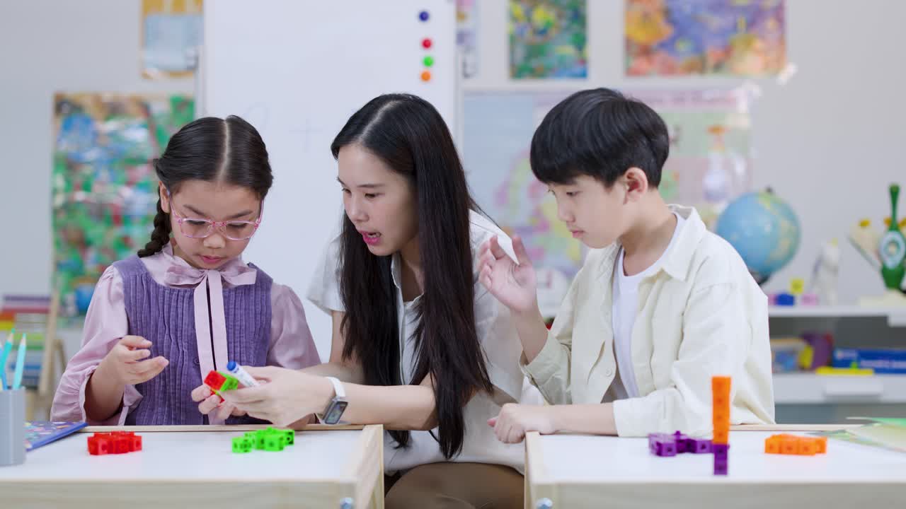 小学校の先生が、カラフルな計算ブロックを使って机で実践的な算数活動をする2人の生徒を指導している様子。