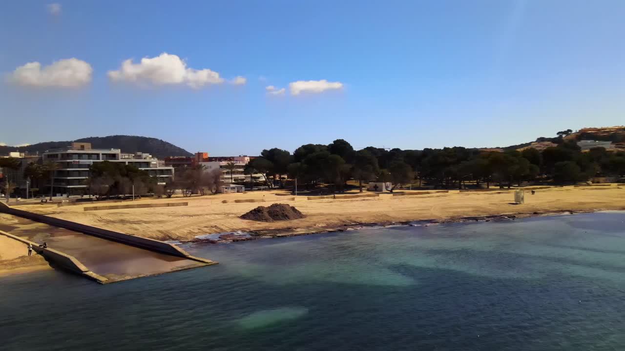 Enjoy a stunning view of the sandy beach in Santa Ponsa, Mallorca. The tranquil waters reflect a bright blue sky while palm trees sway gently in the warm breeze.