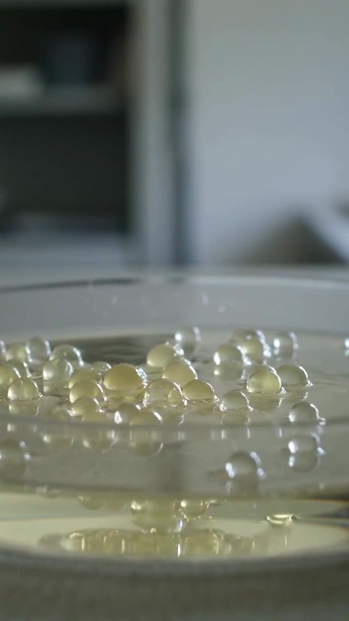 Video verticale: Lievi increspature che muovono perline giallo pallido su liquido trasparente in una capsula di Petri in laboratorio.