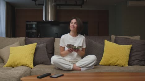 smiling young woman count money cash, financial savings, salary, sitting on couch at home, dreaming about shopping
