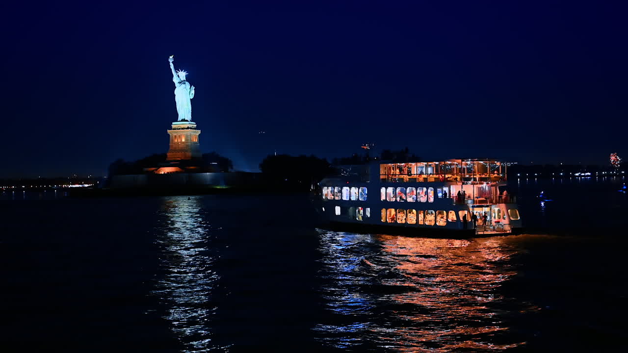 A multi-deck riverboat with switched on illumination travels by the Hudson River. Vehicle approaches the Statue of Liberty well-illuminated at night. River tour by New York, USA