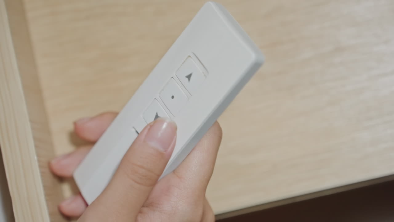 Fjernbetjening ved sengen på hotelværelse, gæst trykker på knapper for at justere persienner og lys på træsengegærde, nærbillede af velplejede fingre, moderne minimal indretning med stille automatiseret komfort.