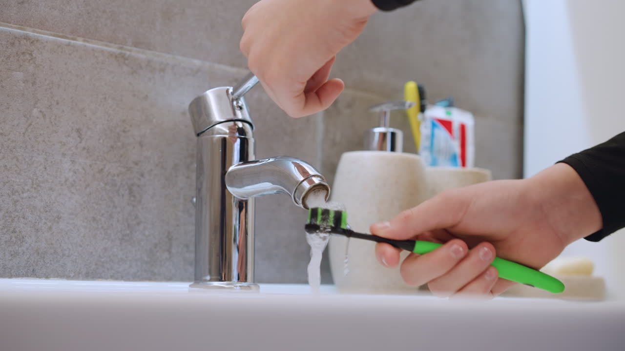 Closeup chrome faucet pouring water over toothbrush in modern bathroom sink, hand steadying green toothbrush, ceramic holders with toothpaste and soap, soft natural light and neutral tile background