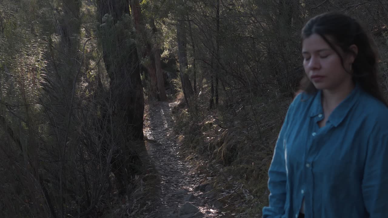 A woman in a blue shirt stands thoughtfully on a forest path surrounded by trees in Warrandyte, Melbourne. The natural setting is peaceful, ideal for introspection
