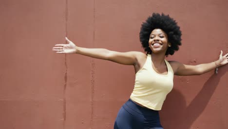 young woman with afro hairstyle wearing sportswear leaning confidently against red wall, radiating happiness with open arms and vibrant smile, embodying joy and urban wellness