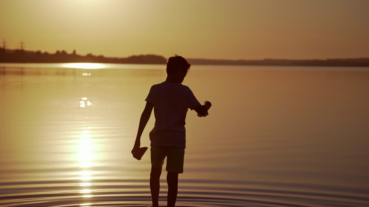 Boy with Red and green Paper Ships is walking in the river. Origami Paper Boats in kid's hands. Beautiful Waves Ripple Background. Travel and nature Concept. Video at sunset.