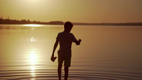 boy with red and green paper ships is walking in the river. origami paper boats in kid's hands. beautiful waves ripple background. travel and nature concept. video at sunset.