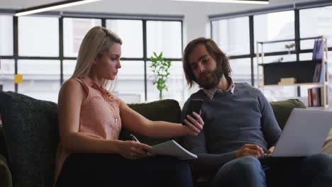 Caucasian-business-people-sitting-on-couch-going-through-paperwork-using-laptop-and-smartphone-in-of