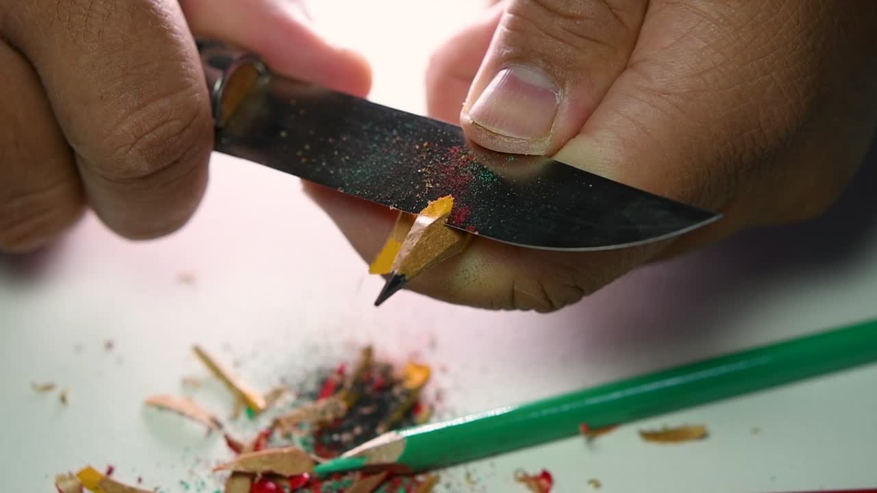 Footage of hands slowly sharpening a pencil and some coloured pencils with a sharp knife