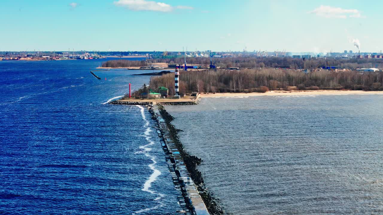 Daugavgrīva Lighthouse and its stone Breakwater at the confluence of the Daugava River and the Baltic Sea, Riga, Latvia, in early spring, under a bright azure sky with sparse white clouds