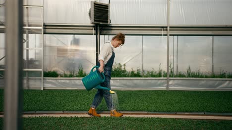 Side-view-of-confident-woman-with-red-hair-farmer-holding-watering-can-in-hands-and-watering-sprouts-of-young-plants-in-greenhouse-on-farm