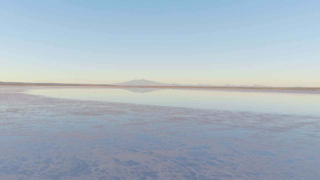 Drone glides smoothly over the vast reflective surface of Salinas del Diamante in southern Mendoza, Argentina, with the faint outline of volcanic peaks and an endless salt horizon under a clear sky