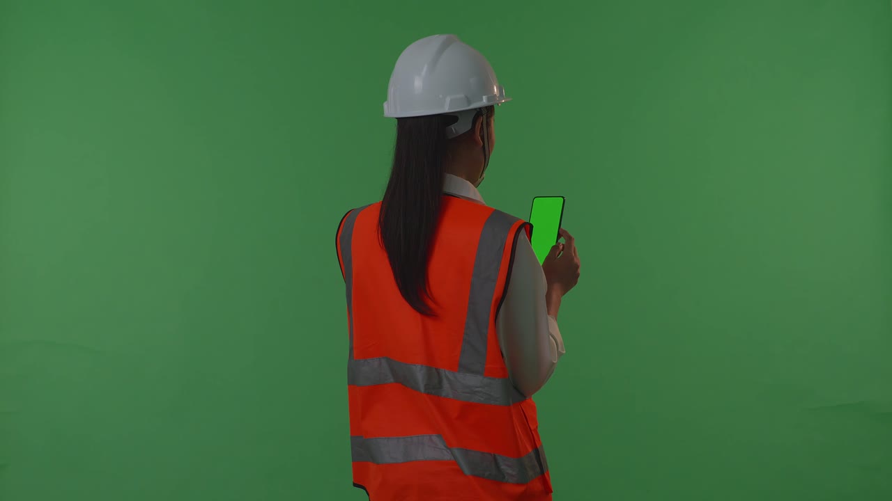 동양 여성 엔지니어가 안전 헬을 착용하여 녹색 스크린 스마트 폰에서 일하는 동안 녹색 화면 배경 스튜디오에 서 있습니다.
