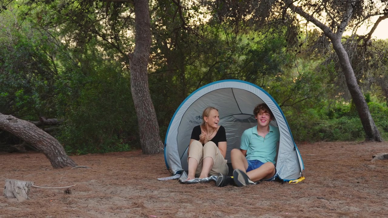 Una madre e suo figlio sono seduti all'interno di una tenda da campeggio, interagiscono e ridono insieme. Sono circondati da alberi e aghi di pino in un ambiente naturale all'aperto