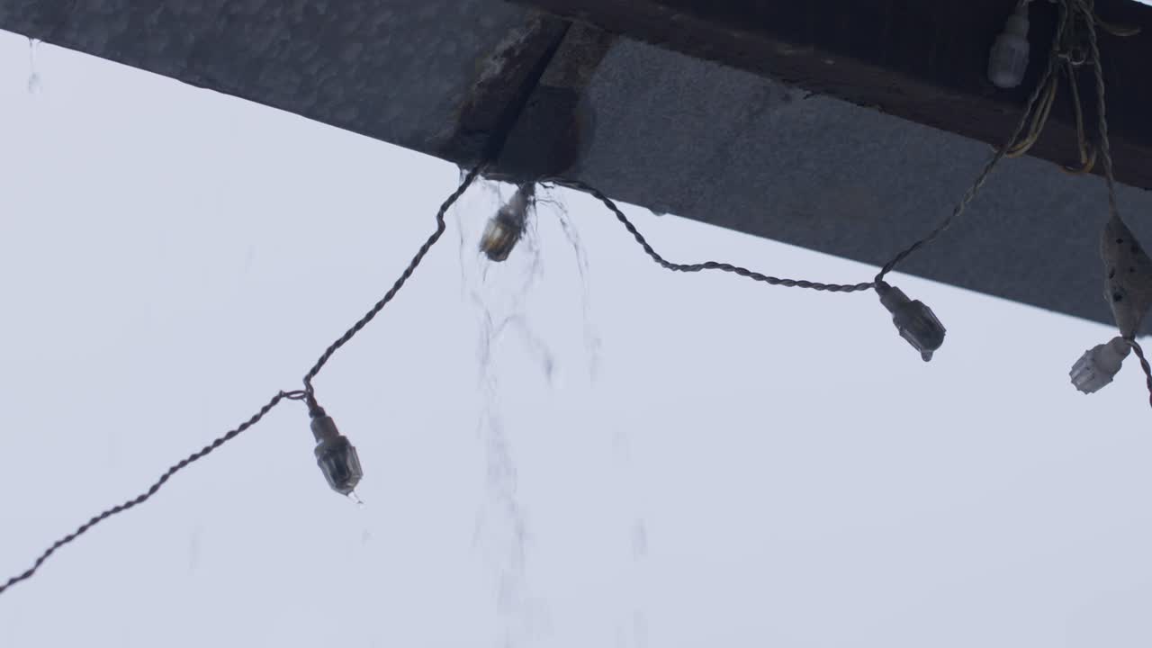 A handheld close up shot of string lights hanging in an outdoor balcony while it rains outside and water flows down on from the light wires