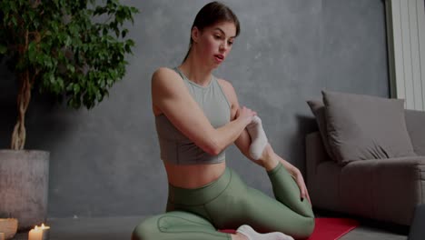 una chica morena confiada en una camiseta deportiva gris y pantalones verdes está involucrada en el estiramiento y el deporte en una alfombra deportiva roja usando lecciones en línea en un apartamento moderno en casa