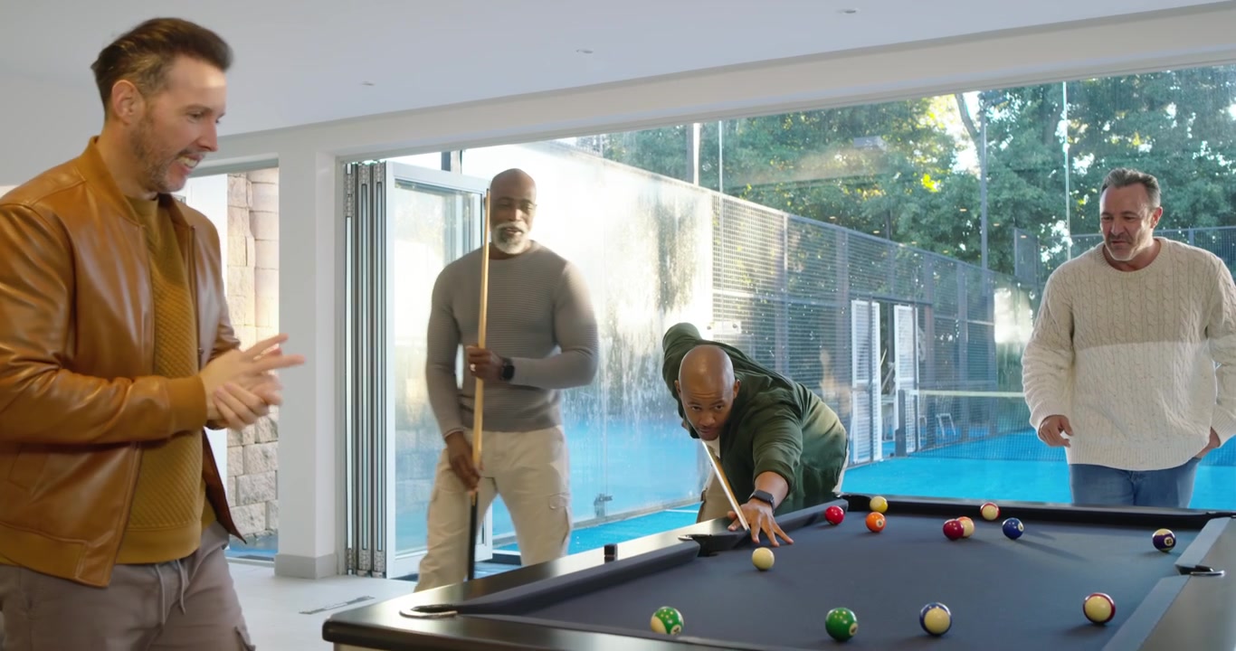Diverse male friends socializing joking urging green top player lining shot at pool near glass wall
