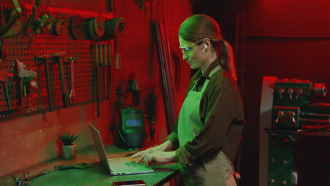 Side-view-of-young-woman-in-apron-and-goggles-using-laptop-in-metal-workshop