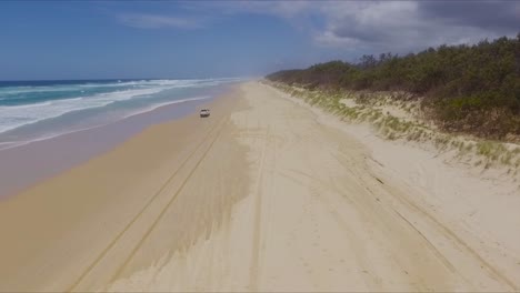 smooth, wide aerial drone shot following a white 4x4 four wheel drive pickup truck over rutted tire tracks, along north stradbroke island's main beach from a distance, as clouds gather over the island