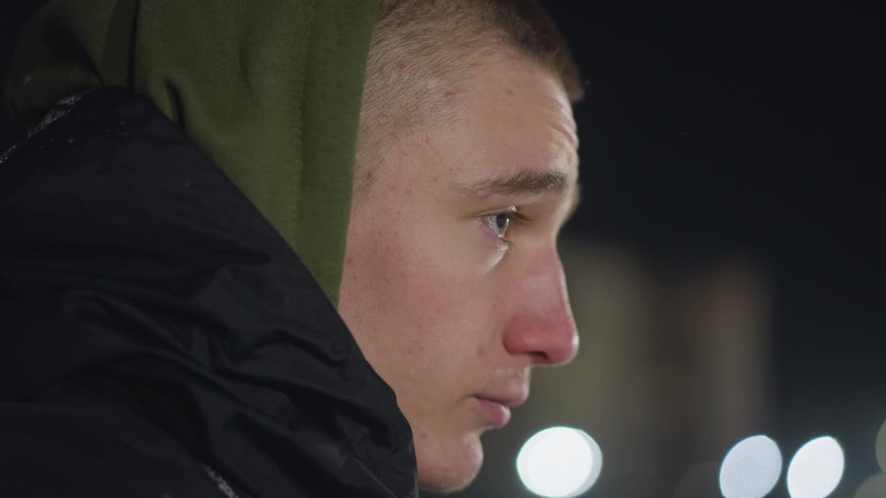 Close up of young man wearing winter hoodie looking around thoughtfully during cold night with soft background blur and city lights creating calm and introspective mood in outdoor setting