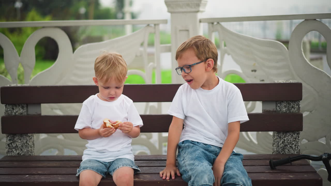 Two siblings are seated on a bench. The younger child offers a piece of his sausage roll to the older brother, who happily accepts and eats it, both are dressed casually